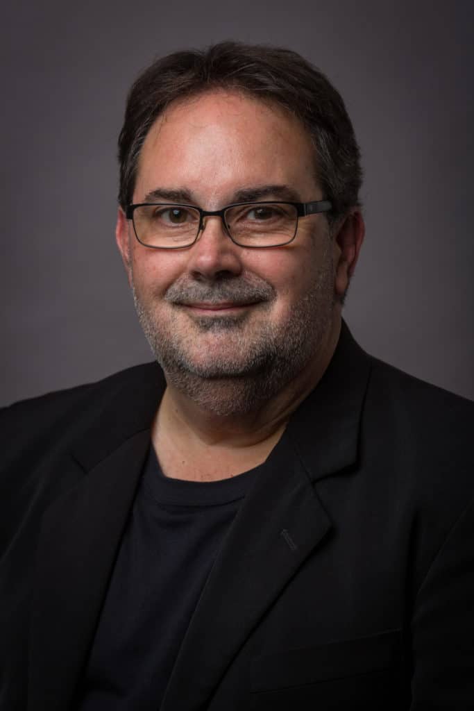 Photo of Brian Nadeau, trumpet instructor, smiling and wearing a black suit jacket and black shirt.