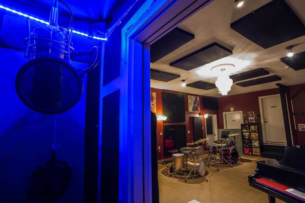 A close up on the recording studio vocal booth looking out into the live room with a drum set and grand piano